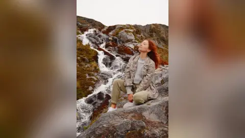LaurentBelluci sits on a wet rock next to a rushing waterfall, wearing a light grey patterned jacket and tan trousers.
