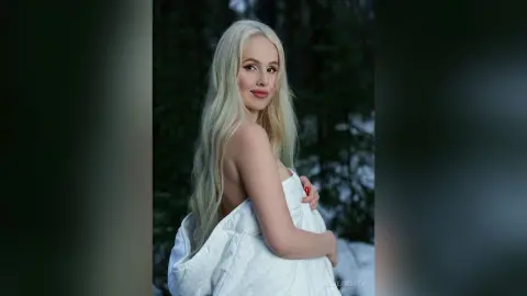 EllieFrosty poses with long blonde hair, wrapped in a white duvet against a dark forest background.