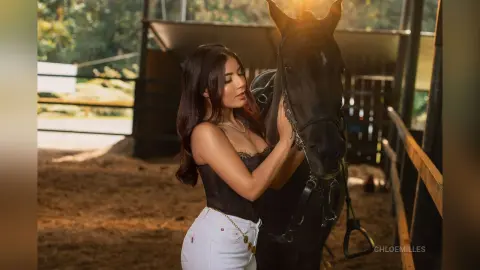 Young woman in a black top and white pants petting a dark horse in a sunlit stable setting.