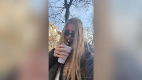 Blonde woman in a black puffer jacket drinking from a cup outdoors on a sunny day.