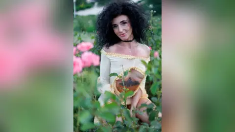 Smiling woman with curly dark hair in a cream lace dress posing in a lush pink rose garden.