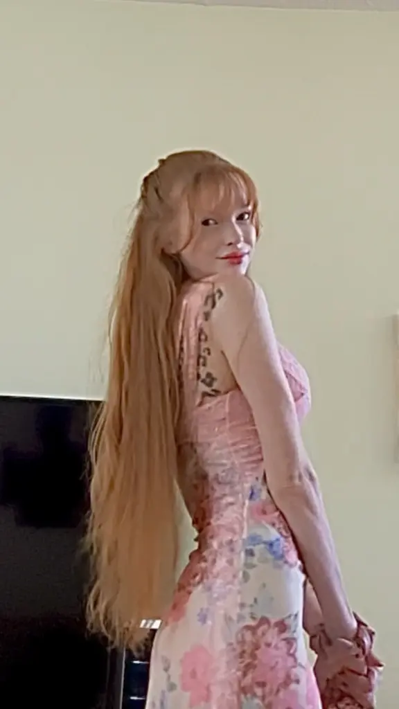 AuroraBlossom poses with her long blonde hair, wearing a pink lace top that shows her shoulders and cleavage.
