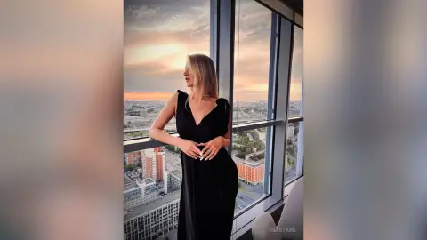InaStark poses in a black dress by a large window overlooking the city at sunset.