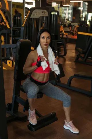 Fit woman in pink sports bra and grey leggings resting with a towel in a modern gym setting.