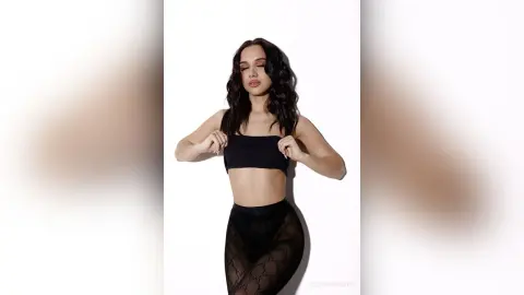 Young woman in black crop top and sheer patterned leggings posing against a white background.