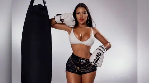 CassandraDuque poses in a white bra and black boxing shorts while wearing white boxing gloves next to a black heavy bag.