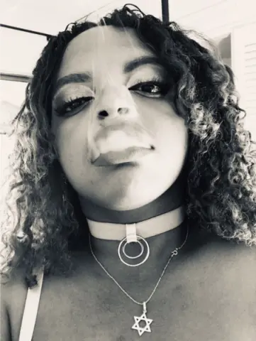 Close-up portrait of a curly-haired woman wearing a choker and Star of David necklace in a moody black and white style.