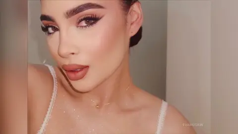 Young woman with glamorous winged eyeliner and glowing skin wearing a white lace camisole in soft indoor lighting.