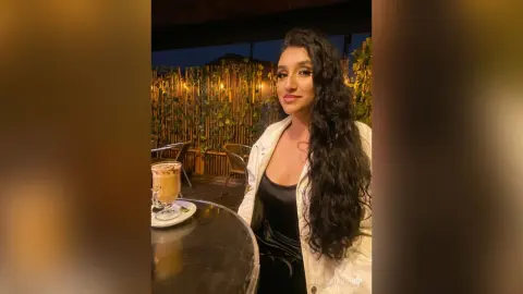 Smiling woman in a white jacket sitting at an outdoor cafe terrace at night.