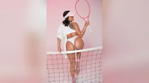 Athletic woman in white tennis outfit and visor holding a pink racket by a net against a pink background.