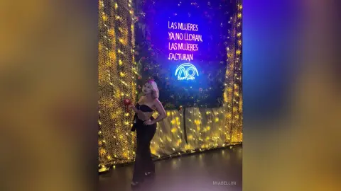 Confident woman in a black outfit holding a red rose against a glowing fairy light backdrop at a festive event.