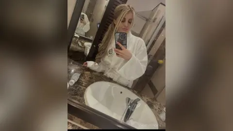 Blonde woman in a white spa bathrobe taking a selfie in a modern marble bathroom mirror.