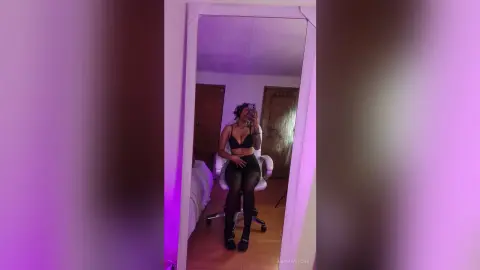 NaomiVelour poses in a black bra and black shorts with sheer black tights, sitting in a white chair in a room with purple lighting.