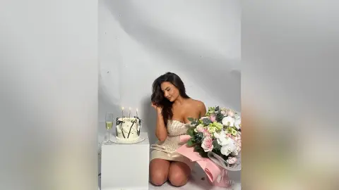 Elegant woman in a gold sequin dress sitting by a birthday cake and pink flower bouquet in a bright studio.