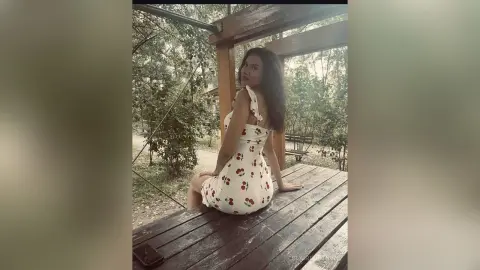 Young woman in a white floral sundress sitting on a wooden deck in a lush, peaceful garden setting.
