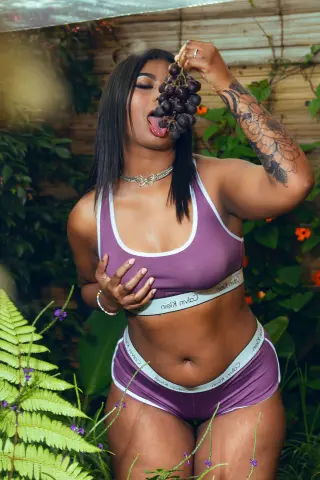 Curvy woman in mauve sports bra and briefs eating grapes in a lush green garden setting.