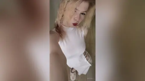 Blonde woman in a white sleeveless top posing with a pouty expression in a modern indoor setting.