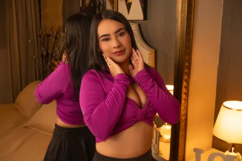 Young woman in a purple long-sleeve crop top posing in a warm, cozy bedroom setting.