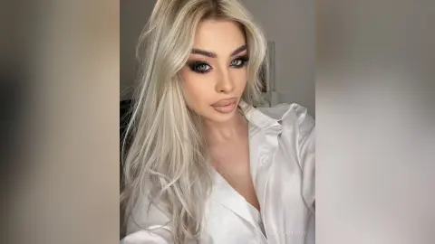 Blonde woman with glamorous smoky eye makeup wearing a white silk blouse in a bright indoor setting.