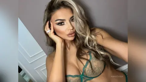 Anastasia poses with her hand against her face, wearing a green lace bra and heavy eye makeup.