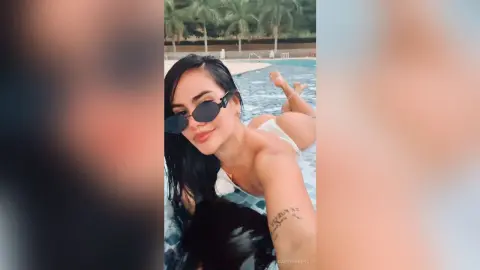 Smiling woman in white swimwear and sunglasses relaxing by a sunny outdoor swimming pool.