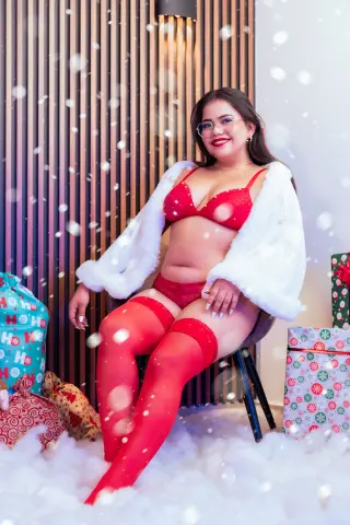 Smiling woman in red lingerie and white faux fur coat sitting amidst festive Christmas decor and snow effects.