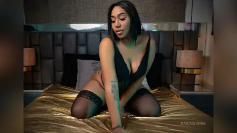 NaiomiLanne — Beautiful Black woman in black lace lingerie and sheer stockings posing on a gold bed in a moody bedroom setting.