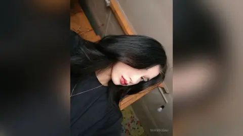 Young woman with long dark hair wearing a black t-shirt in a cozy indoor setting.
