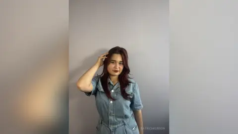 PatriciaGibson poses with her hand in her long dark hair, wearing a light blue denim button-up shirt against a plain grey wall.