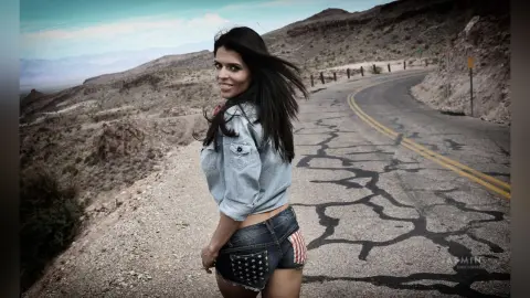 DrellaWolf — Smiling woman in denim jacket and American flag shorts standing on a winding desert mountain road.