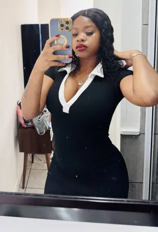 Young woman in a black fitted v-neck dress taking a confident mirror selfie in a bright room.