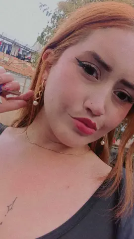 Redheaded woman with winged eyeliner and gold earrings posing outdoors in a casual black tank top.