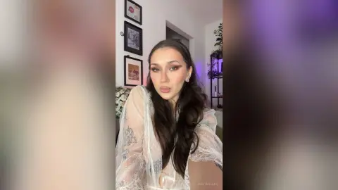 Young woman in a white lace robe posing with soft makeup in a cozy, modern indoor setting.