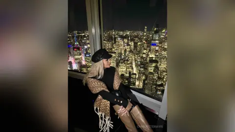 Blonde woman in a black sweater and tan boots posing by a window overlooking a glowing city skyline at night.