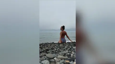 Young woman in a patterned swimsuit sitting on a rocky beach under a misty, moody sky.