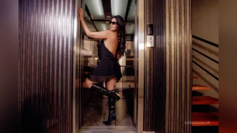Stylish woman in a black mini dress and boots posing confidently in a modern elevator doorway.