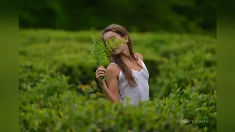 Young woman in a white tank top holding a green fern leaf in a lush, dreamy green field.