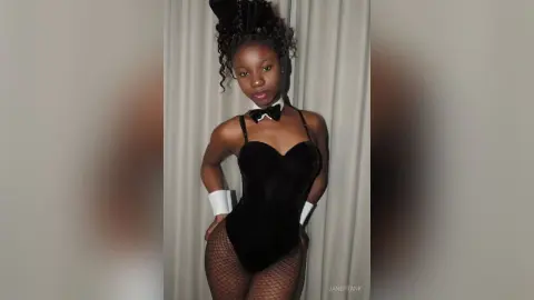 JaneFrank poses in a black bodysuit, bow tie, and fishnet stockings against a white curtain background.