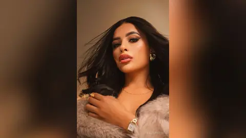 Glamorous woman with long dark hair wearing a fuzzy beige coat and gold jewelry in a warm studio setting.