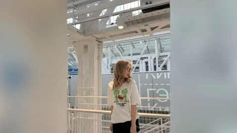 Blonde woman in a white graphic t-shirt posing playfully in a modern white industrial loft setting.