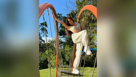 TefyRusso poses outdoors in a white crop top and matching white pants, standing on a hanging bridge with her long brown hair down.