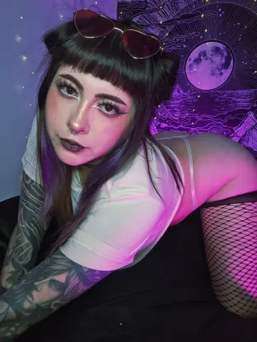 Alt girl with dark bangs and tattoos wearing a white t-shirt and fishnets in moody purple lighting.