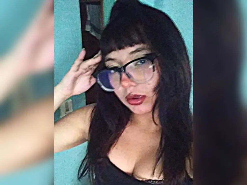 Young woman with dark hair and glasses wearing a black top in a casual indoor setting.