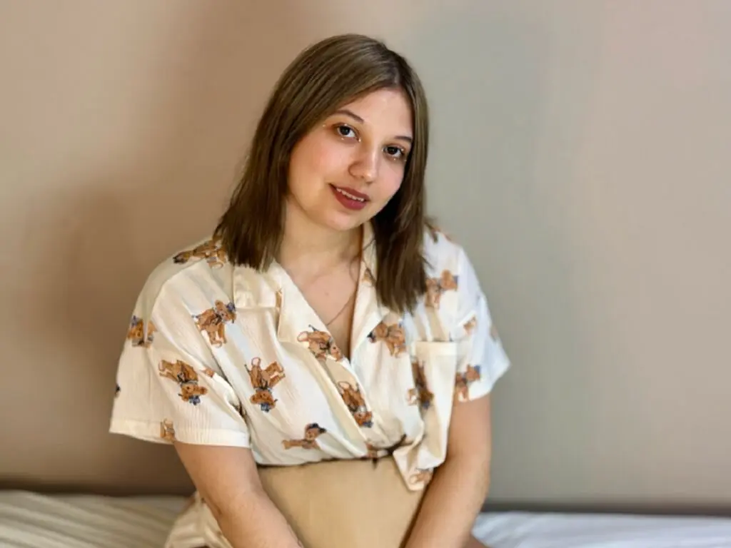 Young woman in a cream patterned short-sleeve shirt sitting relaxed against a neutral wall background.