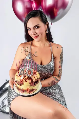 Smiling woman in a silver sequin dress holding a 40th birthday cake with pink balloons in a festive setting.
