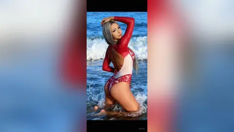 Kalira poses on a beach wearing a red and white lace bodysuit, showing off her long grey hair and bare skin while standing in the ocean waves.