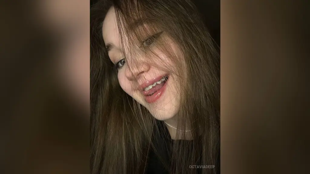 Close-up portrait of a smiling young woman with long brown hair and natural makeup in soft indoor lighting.