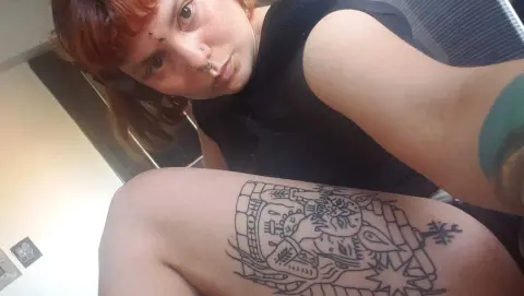 Redheaded woman in a black tank top showing a large black ink line art tattoo on her arm in a bright room.