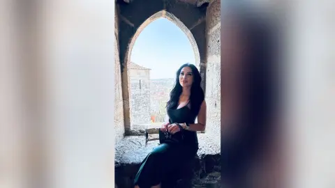 Elegant woman in a black dress sitting by a stone archway overlooking a scenic European cityscape.