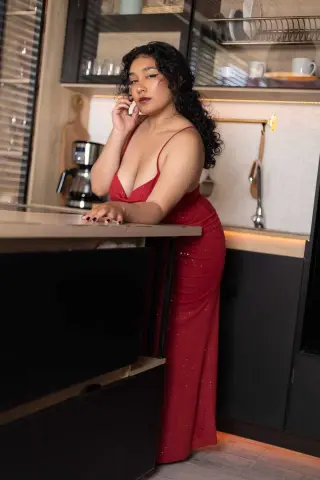 Curvy woman in a glamorous red evening gown posing in a modern kitchen setting.